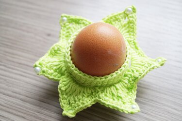 Sternförmiger limettengrüner gehäkelter Eierwärmer mit Perlen um ein braunes Ei auf hellem Holztisch