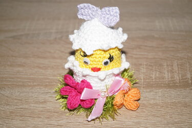 Gelbes gehäkeltes Küken mit Wackelaugen in weißer Eierschale, verziert mit pinken und orangen Blumen sowie rosa und lila Schleifen, auf Holztisch.