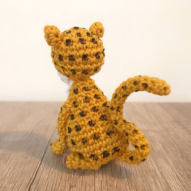 Kleiner gehäkelter Amigurumi-Leopard in Gelb mit schwarzen Flecken, sitzend, von hinten auf Holzuntergrund.