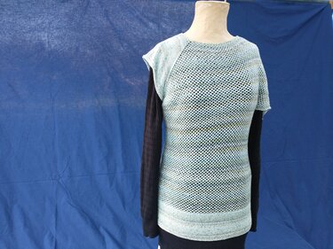 Hellblau-grün meliertes kurzärmliges Strickshirt mit Lochmuster auf Schaufensterpuppe vor blauem Hintergrund