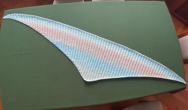 Triangular crochet shawl with pastel pink, blue, and green gradient laid flat on a green surface