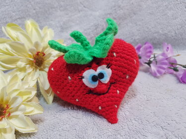 Gehäkeltes rotes Erdbeer‑Herz Amigurumi mit grünen Blättern, weißen Samenstichen und lächelndem Gesicht auf hellem Stoff