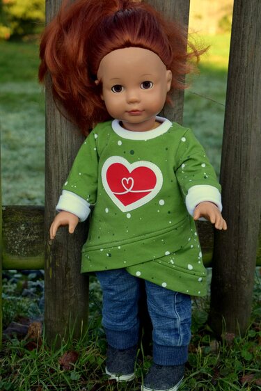 Puppe mit rötlichem Haar trägt grüne Tunika mit rotem Herz und Jeans vor einem Holzzaun.