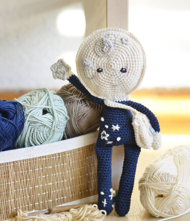 Amigurumi-Mondpuppe in dunkelblauem Outfit neben Korb mit Garn und Häkelhaken