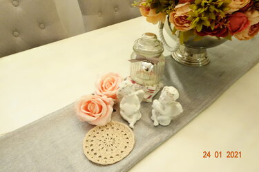 Rundes beiges Häkeldeckchen auf silbernem Tischläufer neben Rosen, Engelsfiguren und Glasdose