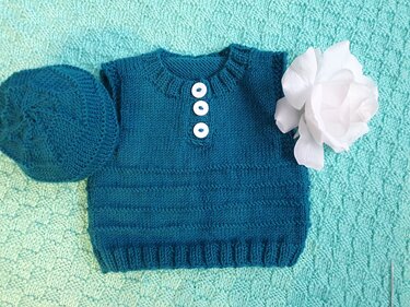 Petrolfarbenes gestricktes Baby-Oberteil mit passender Mütze und drei weißen Knöpfen auf mintgrüner Decke