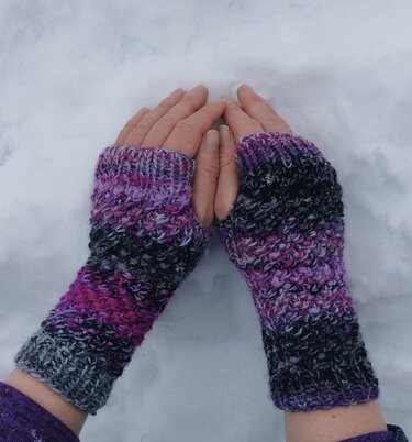 Hände mit fingerlosen, gestrickten Armstulpen in Violett- und Schwarztönen auf Schnee