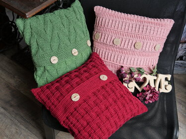 Drei gehäkelte Kissen in Grün, Rosa und Rot mit Holzknöpfen auf einem dunklen Stuhl