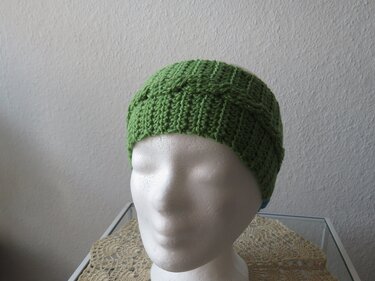 Grünes gehäkeltes Stirnband mit Zopfmuster auf weißem Kopfmodell.