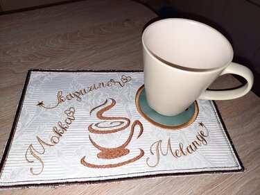 Rechteckiger bestickter Untersetzer mit Kaffeetassen-Motiv und den Schriftzügen 'Mokka', 'Kapuziner', 'Melange', darauf eine weiße Tasse