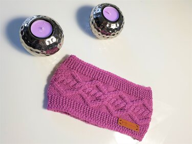 pinkes gestricktes Stirnband mit Zopfmuster und braunem Label, neben zwei silbernen Teelichthaltern
