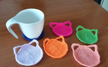 Mehrere kleine gehäkelte Katzen-Untersetzer in verschiedenen Farben auf einem Holztisch