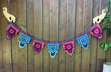 Gehäkelte Wimpelkette mit quadratischen Motiven in Pink, Violett und Blau an einem Holzzaun