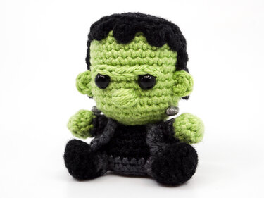 Hand-crocheted green Frankenstein-style amigurumi with black hair, grey neck bolts, and black eyes, sitting on a white background.