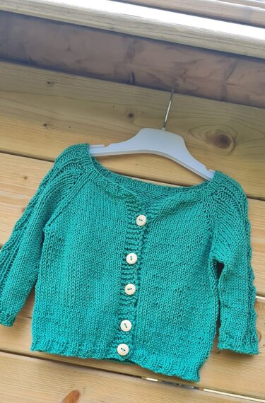 Türkisfarbene gestrickte Babyjacke mit fünf hellen Knöpfen auf einem Kleiderbügel vor einer Holzwand.