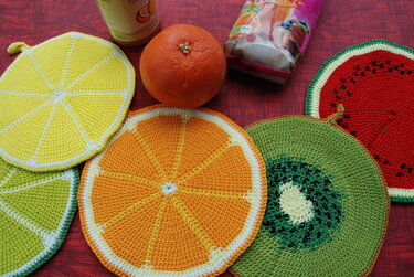 Nahaufnahme von fünf gehäkelten Topflappen in Form von Fruchtscheiben (Zitrone, Orange, Limette, Kiwi, Wassermelone) auf einem roten Tisch.