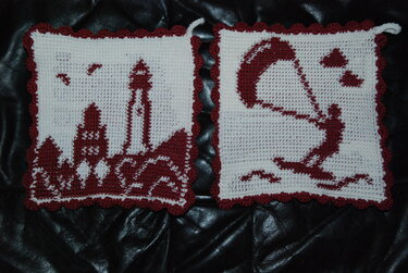 Zwei weiße gehäkelte Topflappen mit dunkelrotem Motiv: links Leuchtturm und Küste, rechts Segel/Windsurfer; gewellter Rand und Aufhängeöse