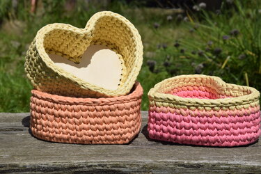 Nahaufnahme von drei kleinen gehäkelten Körben in Pastelltönen: zwei herzförmige und ein runder Korb, auf einer verwitterten Holzbank.