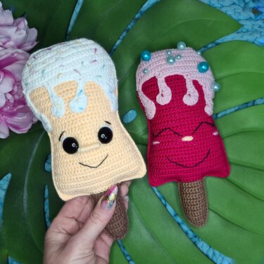 Zwei gehäkelte Eis-Amigurumi: links beige mit weißer Glasur und großen Augen, rechts pink mit Perlen und lächelndem Gesicht; Hand hält das linke Eis