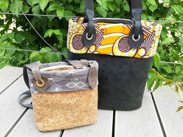 Zwei Taschen auf Holzplanken vor grünem Laub: große Tasche mit schwarzem Kunstlederboden und gelb‑orange gemustertem Stoff, daneben eine kleinere mit Korkboden und grauem, floralem Stoff.