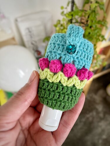 Hand hält kleines gehäkeltes Täschchen in Türkis, Pink, Hellgrün und Grün mit Knopfverschluss und kleiner Reise-Flasche