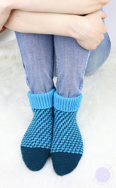 Feet wearing two-tone blue crocheted slippers with folded ribbed cuffs, resting on a white fluffy rug.