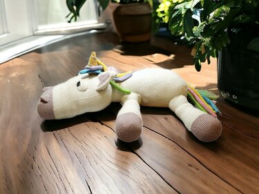 Cream crocheted amigurumi unicorn with pastel rainbow mane and tail on a wooden surface.