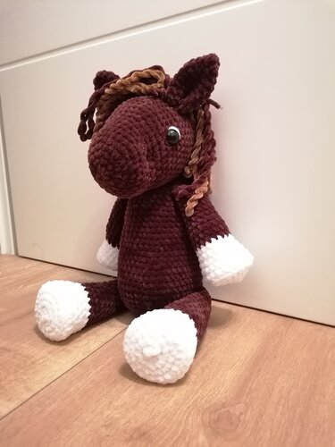 Gehäkeltes dunkelbraunes Amigurumi-Pony mit weißen Hufen und hellbrauner Mähne, sitzend auf Holzboden