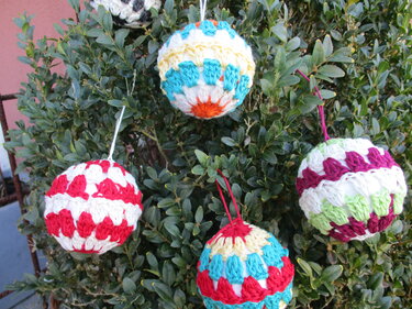 Fünf gehäkelte Weihnachtskugeln mit bunten Streifen und Muschelmuster an einem grünen Strauch