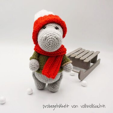 Gehäkeltes graues Amigurumi-Nilpferd mit roter Mütze und Schal neben kleinem Holzschlitten auf weißem Hintergrund.