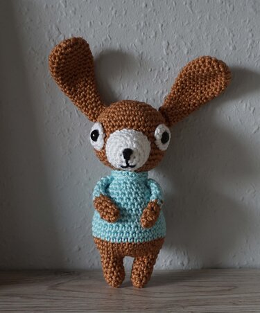 Brown crocheted amigurumi rabbit wearing a light blue sweater with long ears standing on a wooden surface
