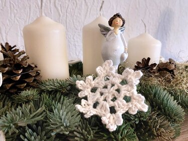 Weiße, schnell gehäkelte Schneeflocke auf einem Adventskranz aus Tannengrün neben Kerzen und Zapfen