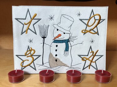 Bestickter Schneemann mit Zylinder, blauem Schal und vier Sternen mit den Zahlen 1, 2, 3, 4; davor vier rote Teelichter.