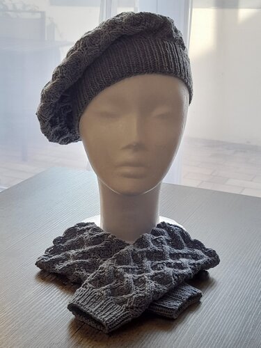 Graue gestrickte Baskenmütze auf Schaufensterkopf und passende fingerlose Handschuhe mit Strukturmuster auf einem Tisch.