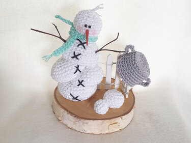 Gehäkelter weißer Amigurumi‑Schneemann mit türkisfarbenem Schal auf Birkenholzscheibe