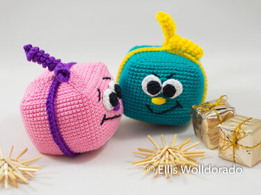 Zwei gehäkelte Geschenkfiguren in Pink und Türkis mit aufgenähten Augen, daneben goldene Päckchen und Strohsterne auf hellem Hintergrund.