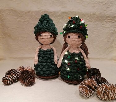 Zwei gehäkelte Amigurumi-Tannenbaum-Puppen, eine mit Lichterkette, umgeben von Kiefernzapfen.