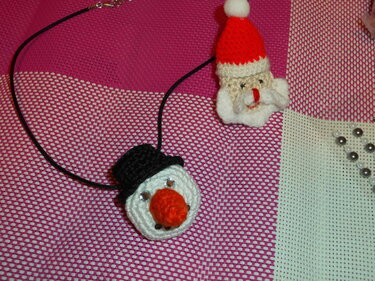 Gehäkelter Schneemann-Anhänger mit schwarzem Hut und oranger Nase an schwarzer Kordelkette neben einer kleinen Weihnachtsmann-Figur auf pink-weißem Stoff.