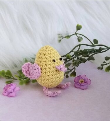 Gelbes Amigurumi-Küken mit rosa Flügeln, Schnabel und Füßen vor hellem Hintergrund, dekoriert mit grünen Zweigen und kleinen Blüten.