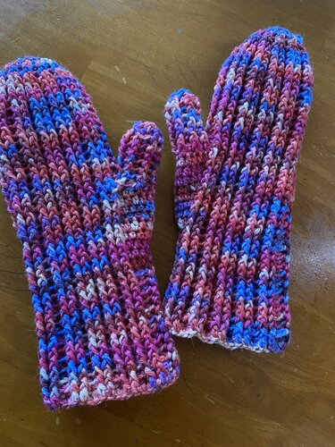 Zwei bunte gehäkelte Fäustlinge in Pink, Lila und Blau auf einem Holztisch.