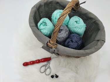 Korb mit Garnknäueln, Häkelnadel, Schere und zwei Sicherheitsaugen auf hellem Untergrund