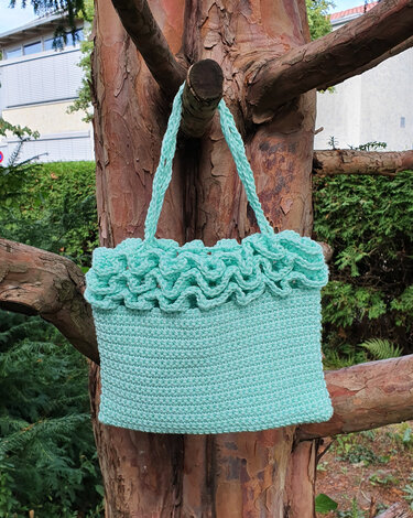 Gehäkelte mintfarbene Henkeltasche mit Rüschenrand und gedrehten Trägern, an einem Baum aufgehängt