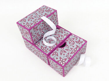 Drei rechteckige Geschenkboxen mit floralem Muster in Grau, Magenta und Weiß, mit weißem Band auf hellem Hintergrund.