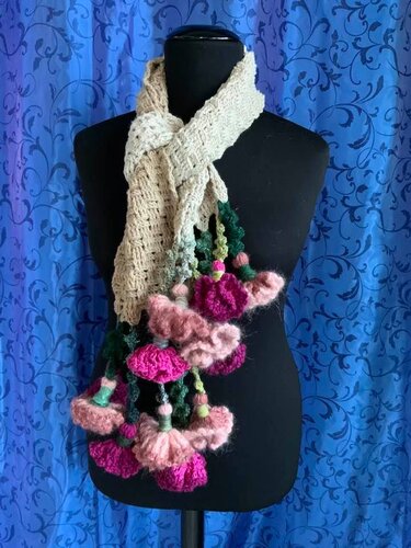 Heller gehäkelter Schal mit rosa und pinken Blütenanhängern auf schwarzer Schneiderpuppe vor blauem Hintergrund