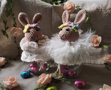 Zwei gehäkelte Hasenköpfe mit Blumenkränzen als dekorative Glastopper auf Gläsern, umgeben von Schokoladeneiern und Rosen