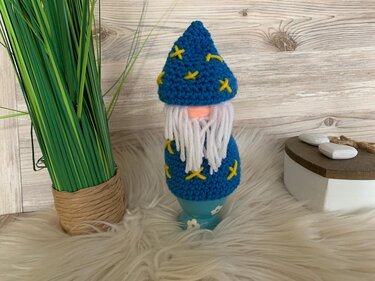 Blauer handgehäkelter Gnom-Eierwärmer mit gelben Sternen, weißem Bart und rosa Nase auf einem hellblauen Eierbecher vor Holzhintergrund.
