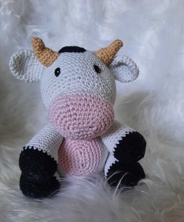 Gehäkelte Amigurumi-Kuh, weiß mit rosa Schnauze und schwarzen Hufen, sitzend