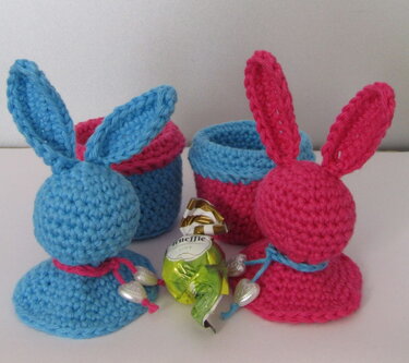 Zwei gehäkelte Hasen-Eierwärmer in Blau und Pink mit kleinen Körbchen und Praline