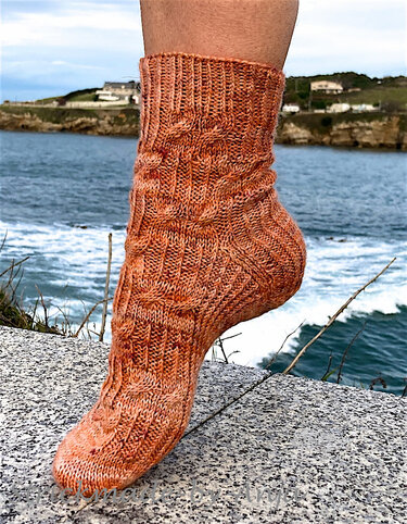 Seitliche Nahaufnahme einer orange-braunen handgestrickten Damensocke mit Zopf- und Rippenmustern, vor Küstenhintergrund