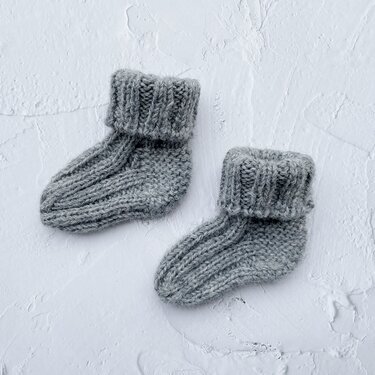 Zwei gestrickte graue Babysocken mit umgeschlagenen Bündchen auf hellem, strukturiertem Untergrund.